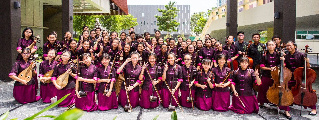 A large group of students in matching dark purple and pink uniforms pose outdoors holding Chinese musical instruments.